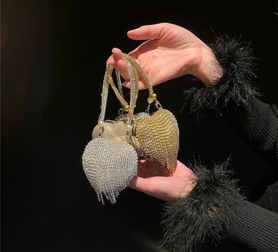 Hand holding two heart-shaped mini chain bags with crystal fringe detailing, one in gold and one in silver, elegant evening accessory for women
