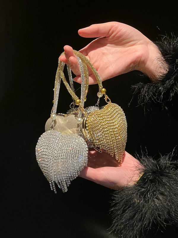 Hand holding two heart-shaped mini chain bags with crystal fringe detailing, one in gold and one in silver, elegant evening accessory for women