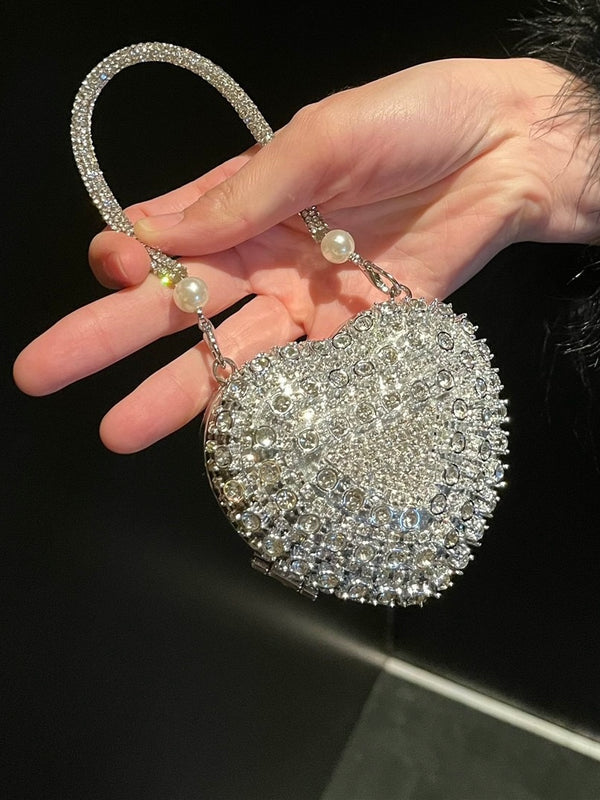 Hand holding a sparkling heart mini handbag with pearl accents luxe against a dark background